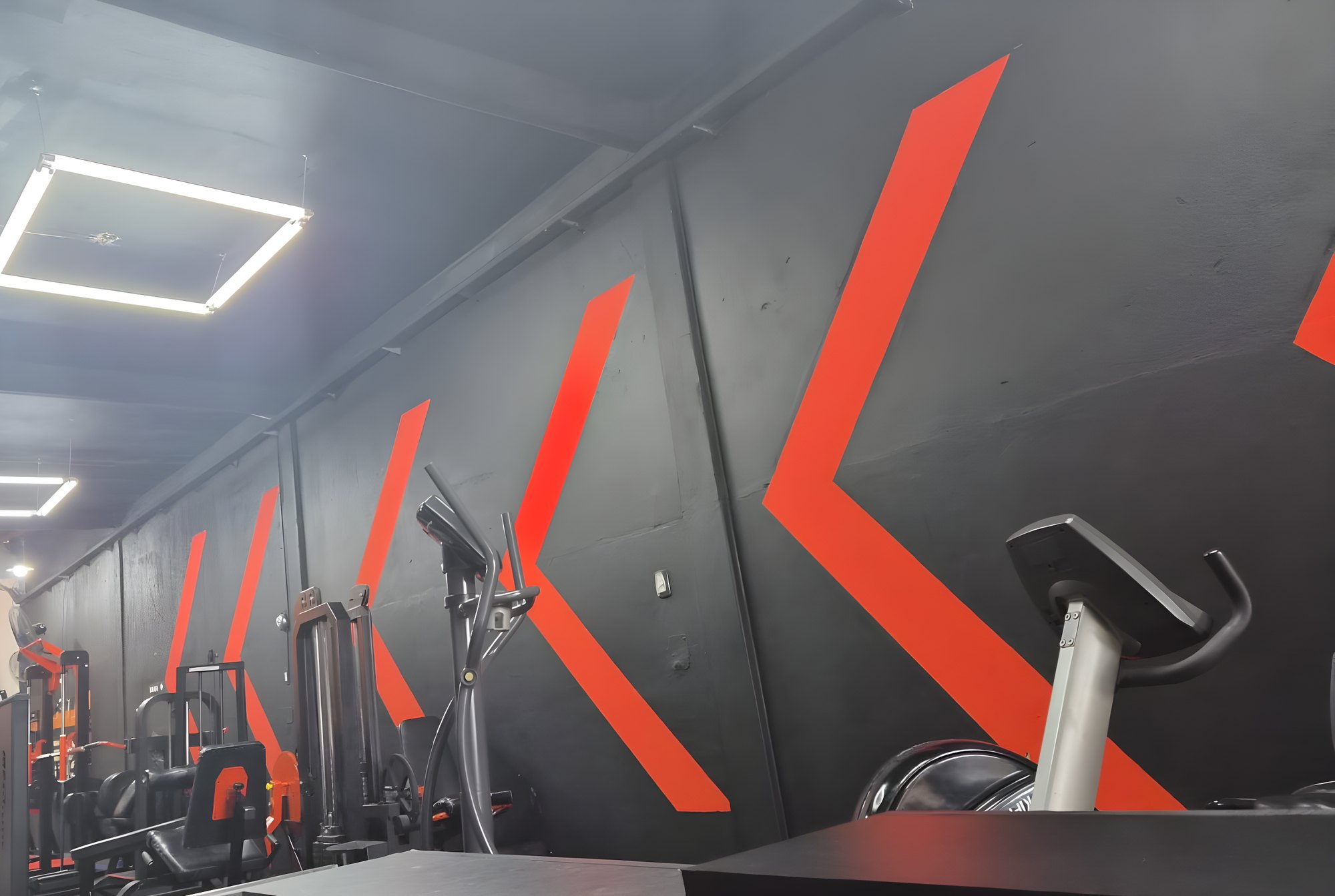Sala de musculacion de Key Fit Gym: pared negra con chevrones naranjas, maquinas de poleas y bicicleta fija