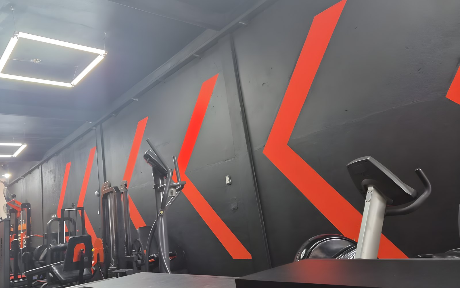 Sector de musculacion de Key Fit Gym en agosto de 2026: pared negra con chevrones naranjas y maquinas de poleas